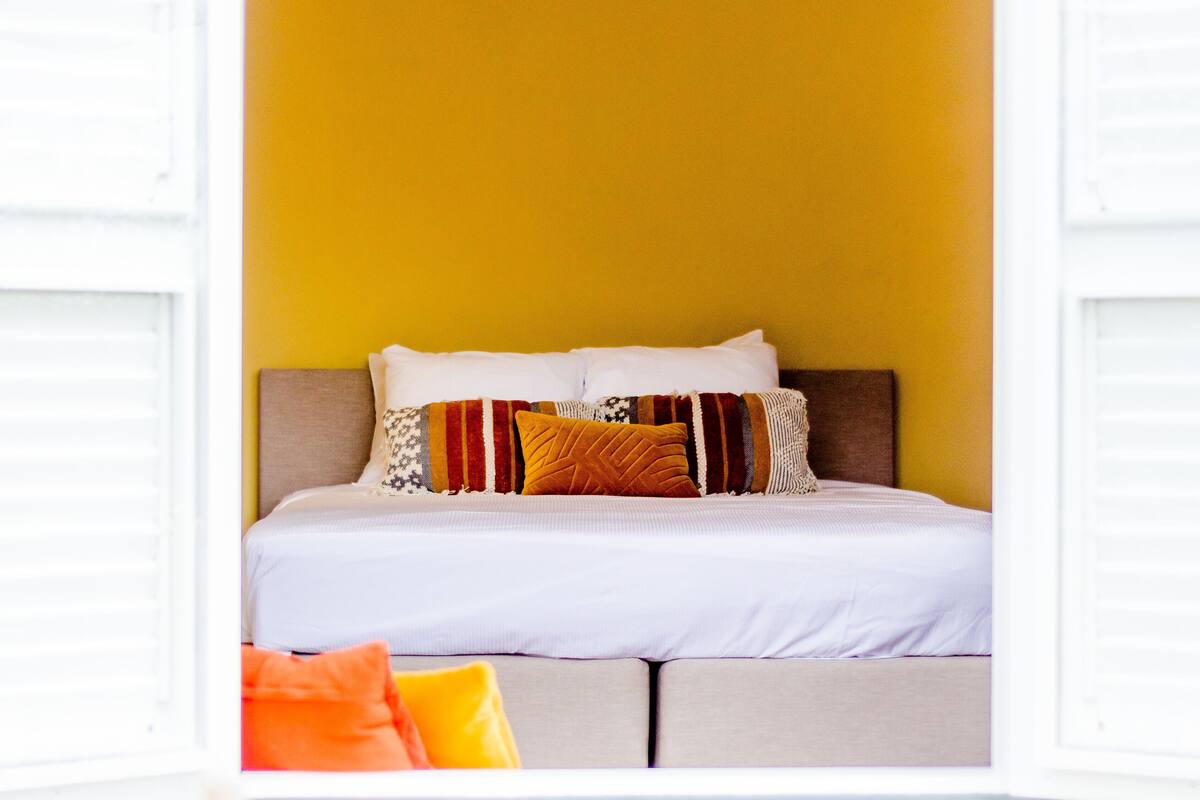 Een bed met witte beddengoed, meerdere kussens en een hoofdbord tegen een gele muur.