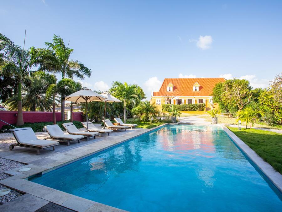 A swimming pool with lounge chairs, a large house with a red roof, and palm trees.
