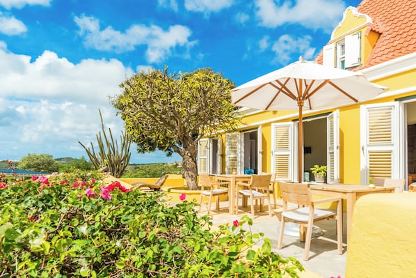 An outdoor seating area with wooden tables and chairs, a white umbrella, and a view of the sea. - Plantation Jan Thiel Lodge (Jan Thiel)
