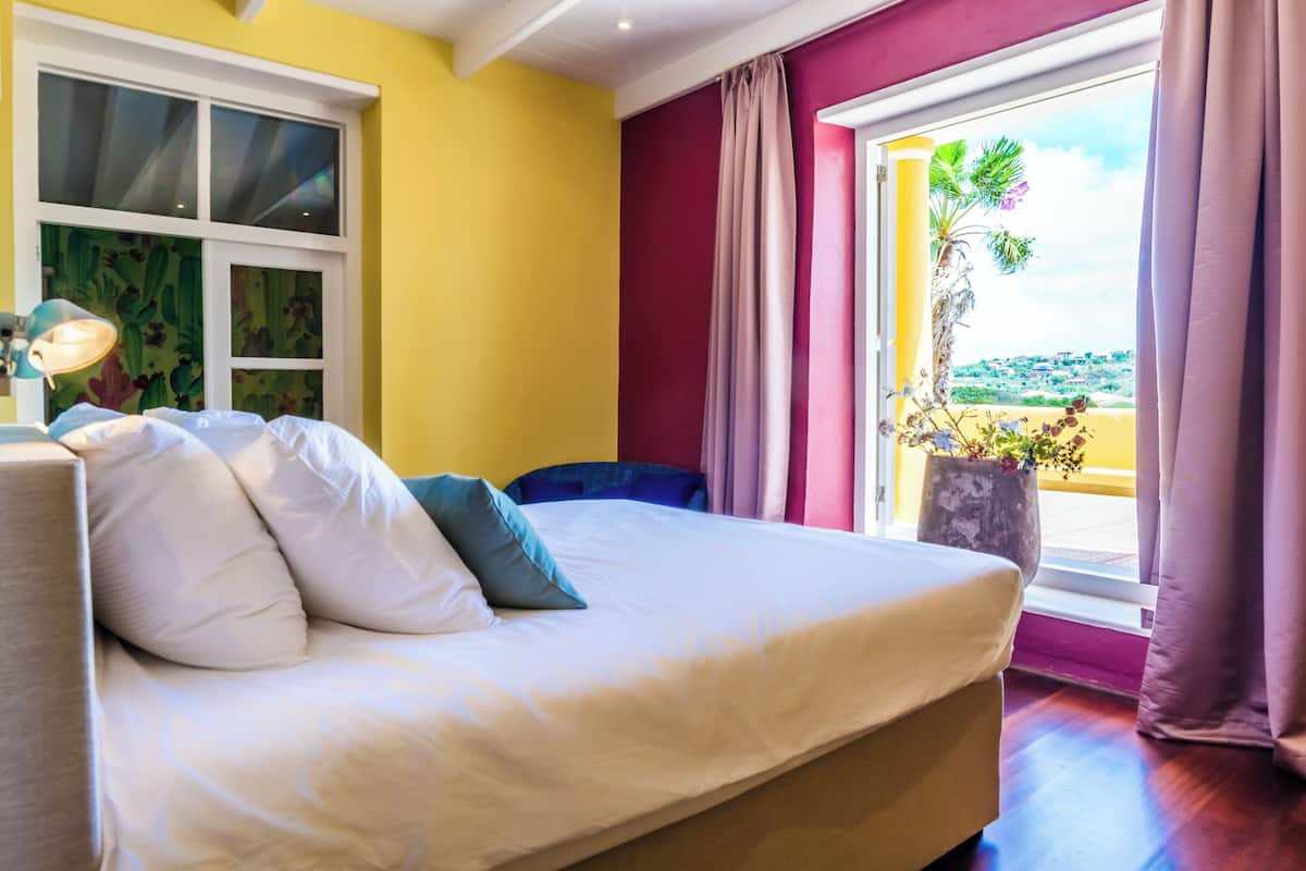Een bed met witte en blauwe kussens, een nachtkastje met een lamp, een raam met een cactusschildering en uitzicht op een palmboom en landschap.