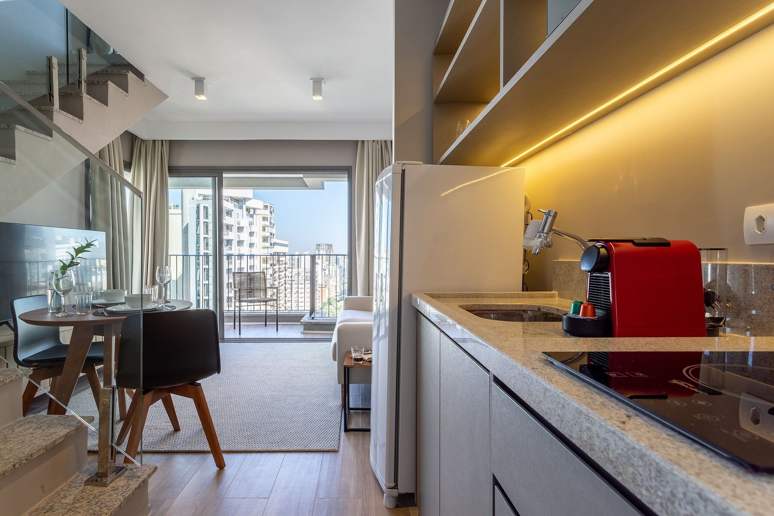 A modern kitchen with a built-in espresso machine, a dining area with a table and chairs, and a view of the city through large windows.