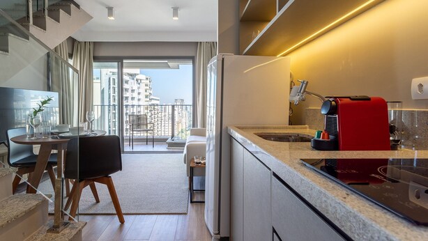A modern kitchen with a built-in espresso machine, a dining area with a table and chairs, and a view of the city through large windows. - Next Haddock by Viva - Apartamentos próximos ao MASP e a metros da Estação Consolação e Avenida Paulista (São Paulo)