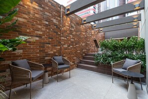 An outdoor seating area with wicker chairs and a tiled floor, a brick wall, and a staircase leading to an upper level. - Next Haddock by Viva - Apartamentos próximos ao MASP e a metros da Estação Consolação e Avenida Paulista (São Paulo)
