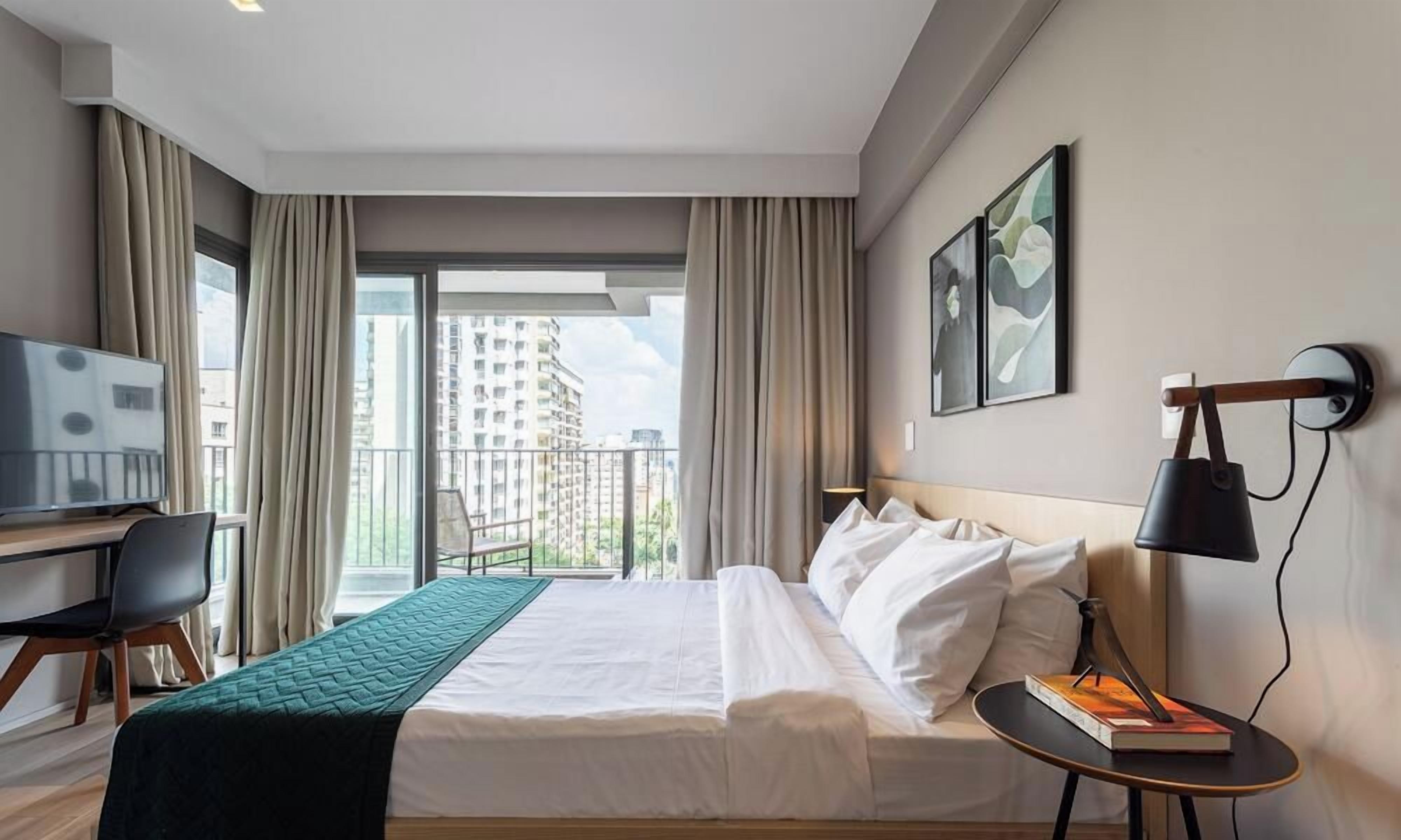 A hotel room with a bed, a desk, a chair, a TV, a balcony with curtains, and artwork on the wall.