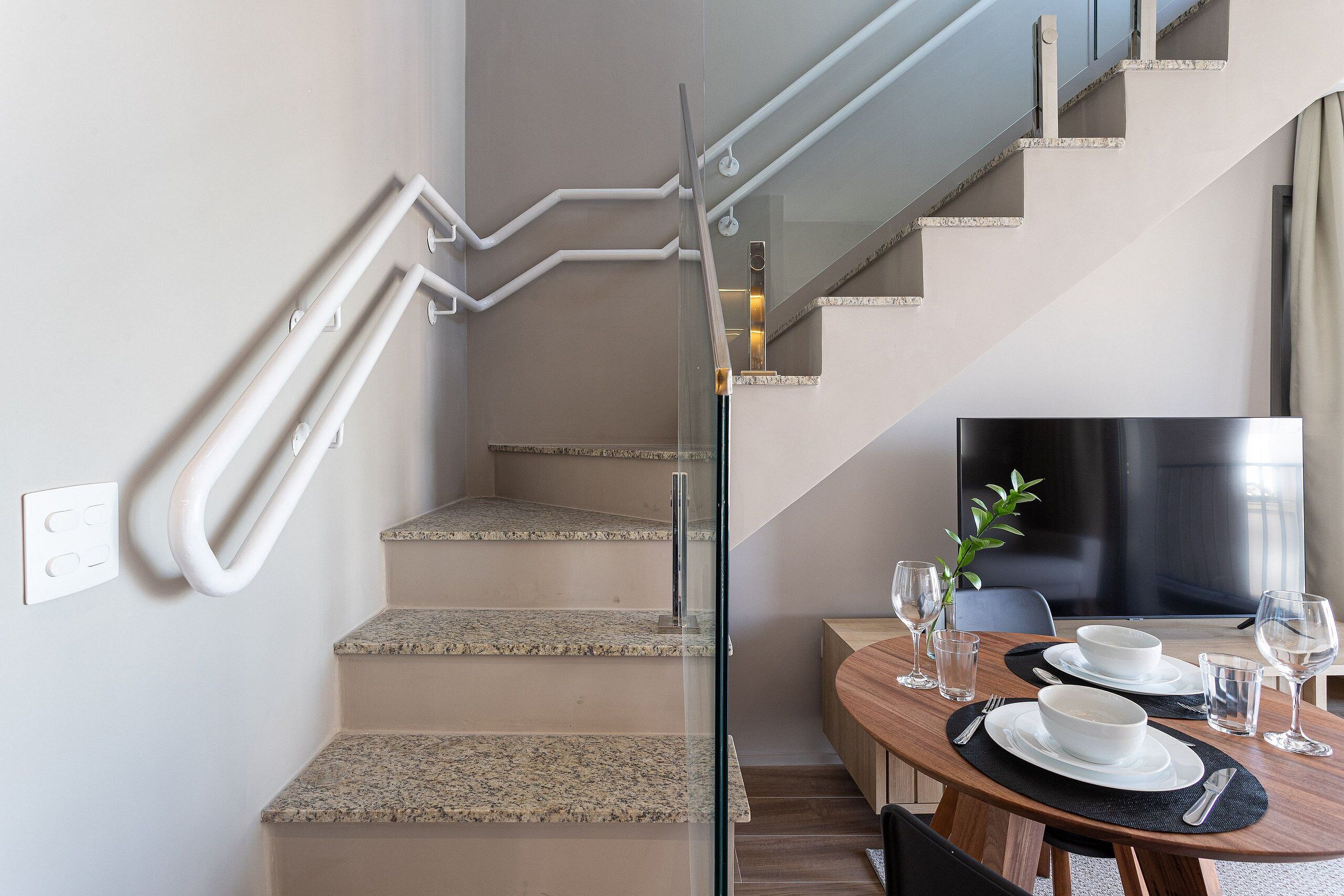 A modern interior with a staircase, a dining area with a table set for two, and a large flat-screen TV.