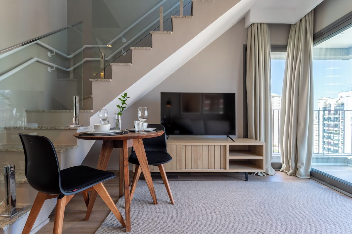 Ein modernes Wohnzimmer mit Fernseher, einem Esstisch mit Stühlen, einer Treppe und einem großen Fenster mit Vorhängen.