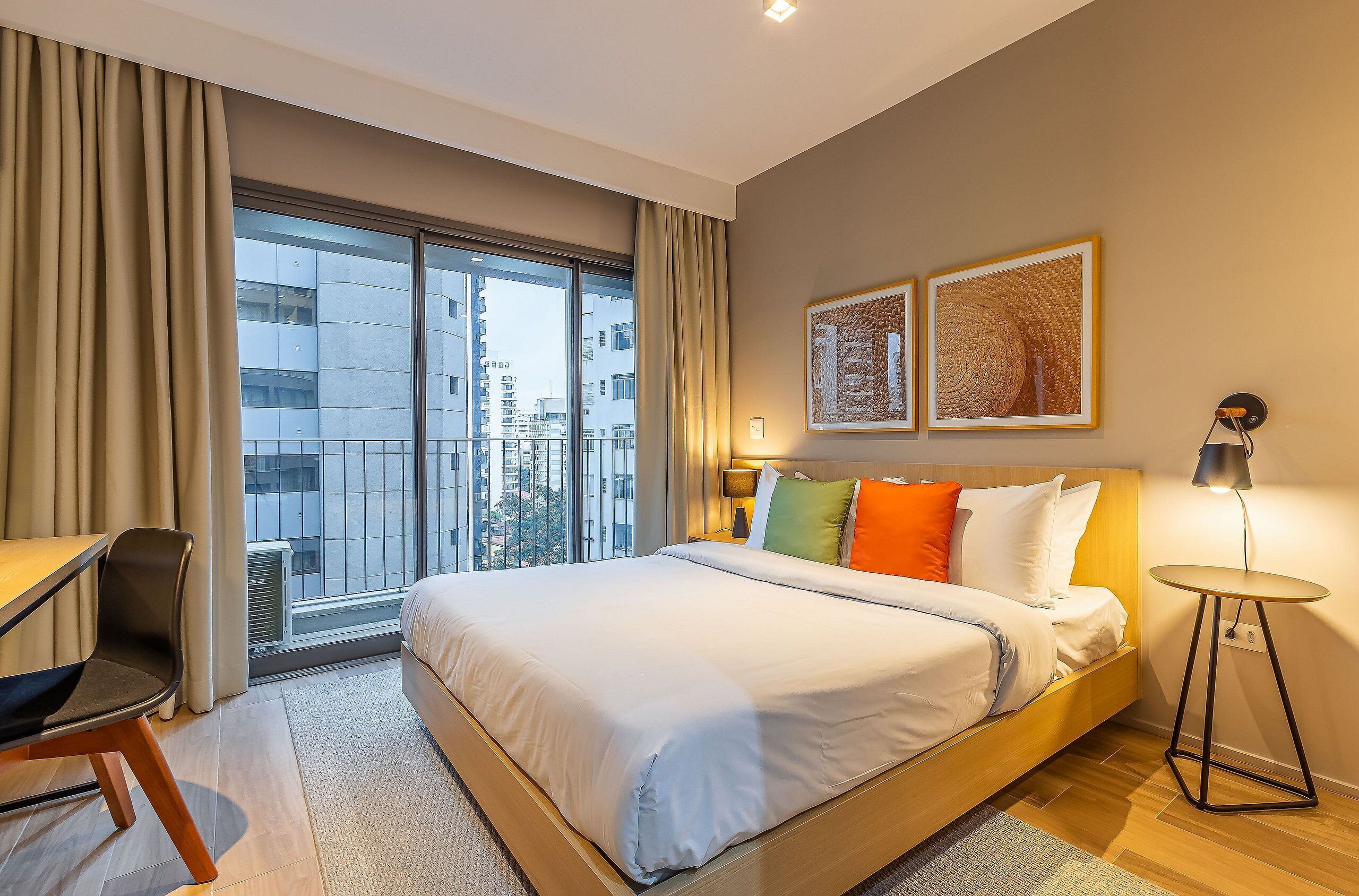 A hotel room with a large bed, a desk, a chair, a small table, and a balcony with a view of the city.