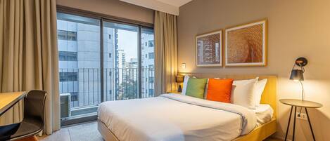A hotel room with a large bed, a desk, a chair, a small table, and a balcony with a view of the city.