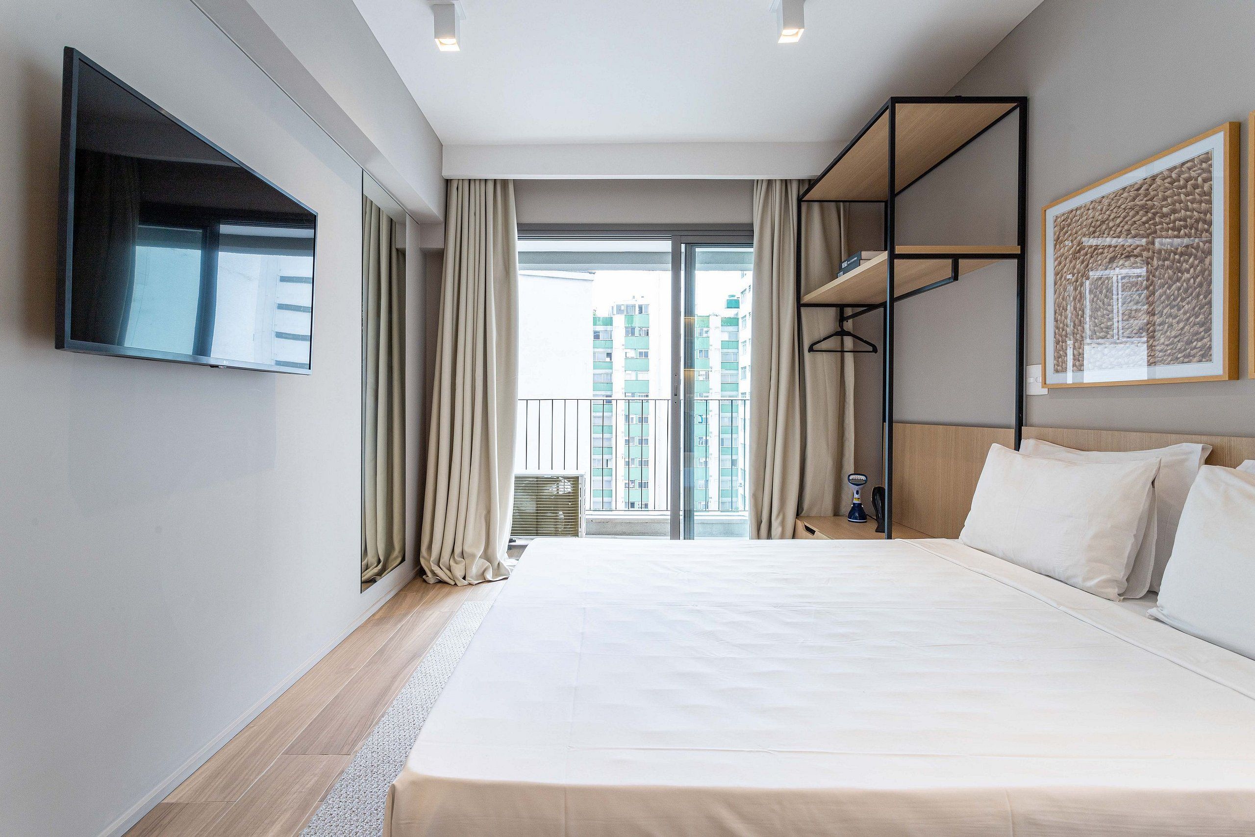 Ein Hotelzimmer mit einem großen Bett, einem Wand-Fernseher, einem Balkon mit Vorhängen und Blick auf Gebäude.