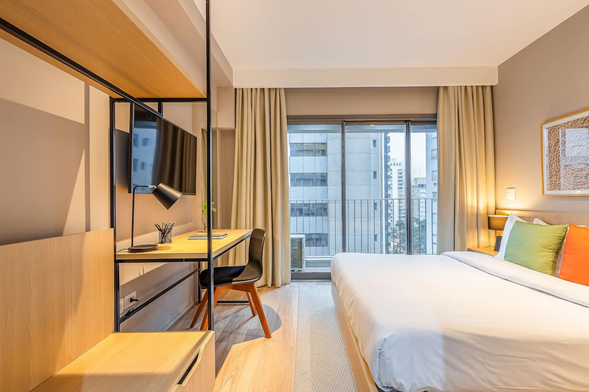 Ein Hotelzimmer mit Bett, Schreibtisch, Stuhl, Fernseher und Blick auf die Stadt durch das Fenster.
