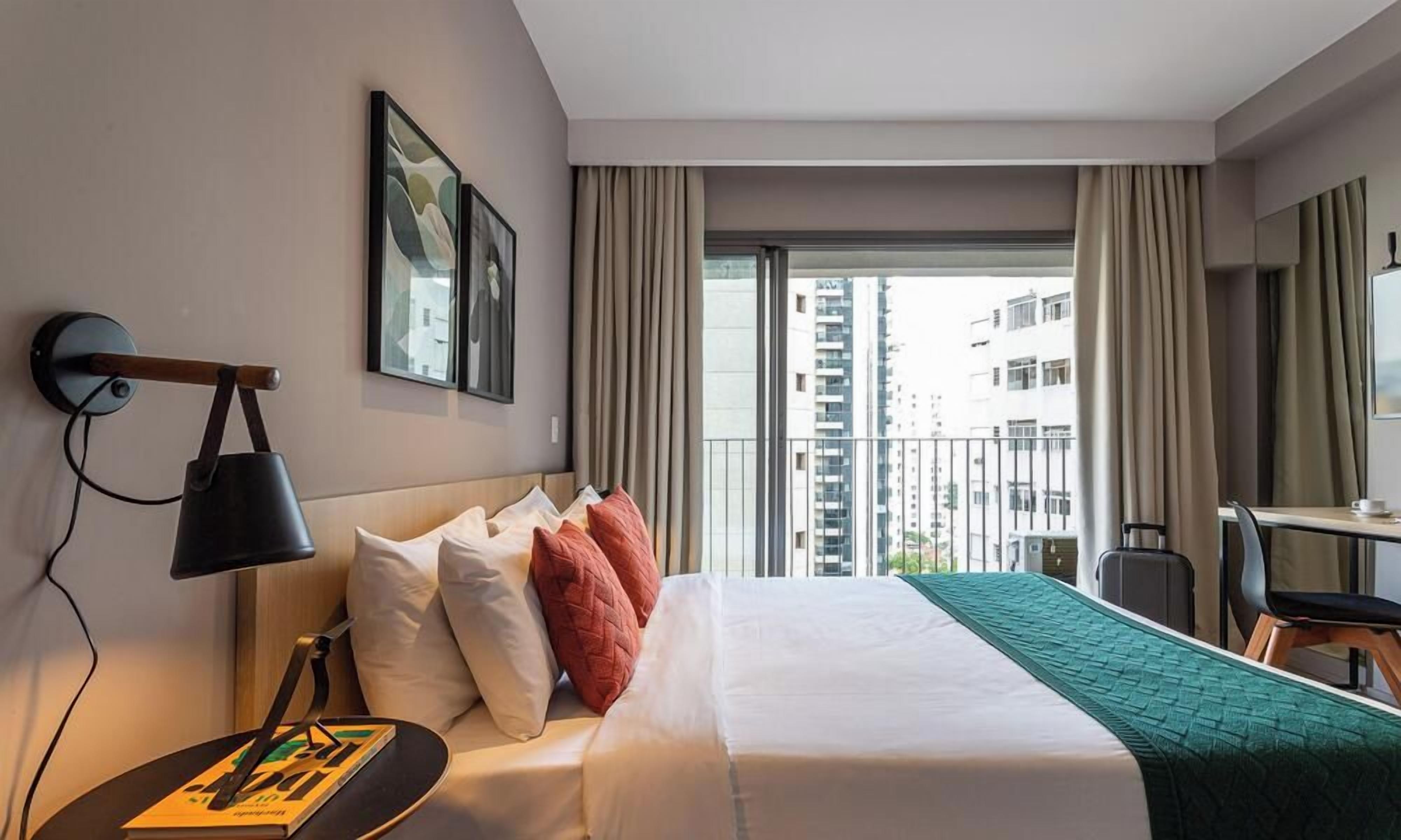 A hotel room with a bed, a desk, a chair, a lamp, and a window with curtains.