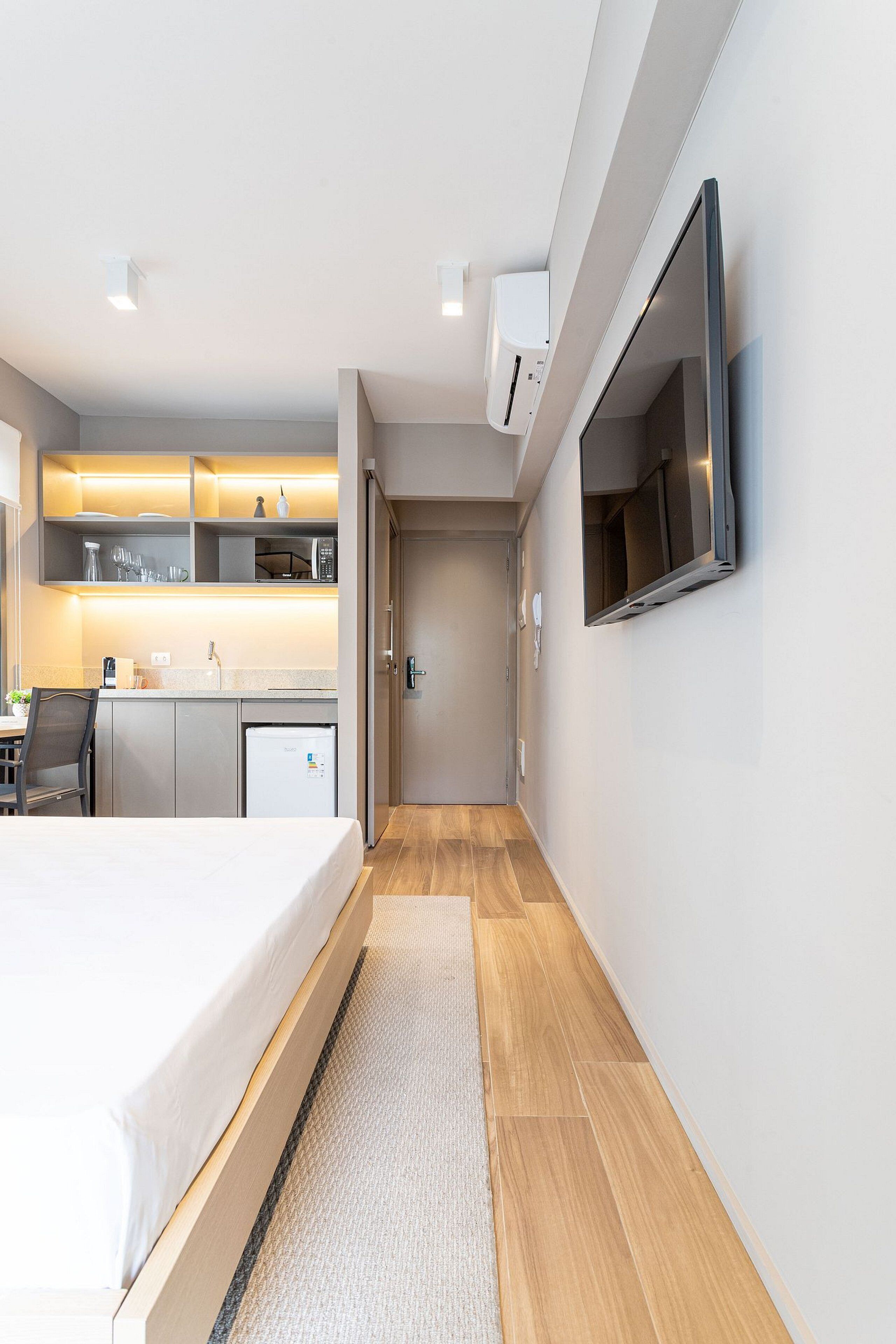 A modern hotel room with a bed, a small kitchenette, a flat-screen TV mounted on the wall, and a wooden floor.