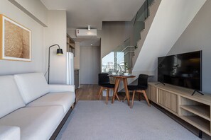 A modern living space with a sofa, a dining table with chairs, a flat-screen TV, and a staircase leading to an upper level.