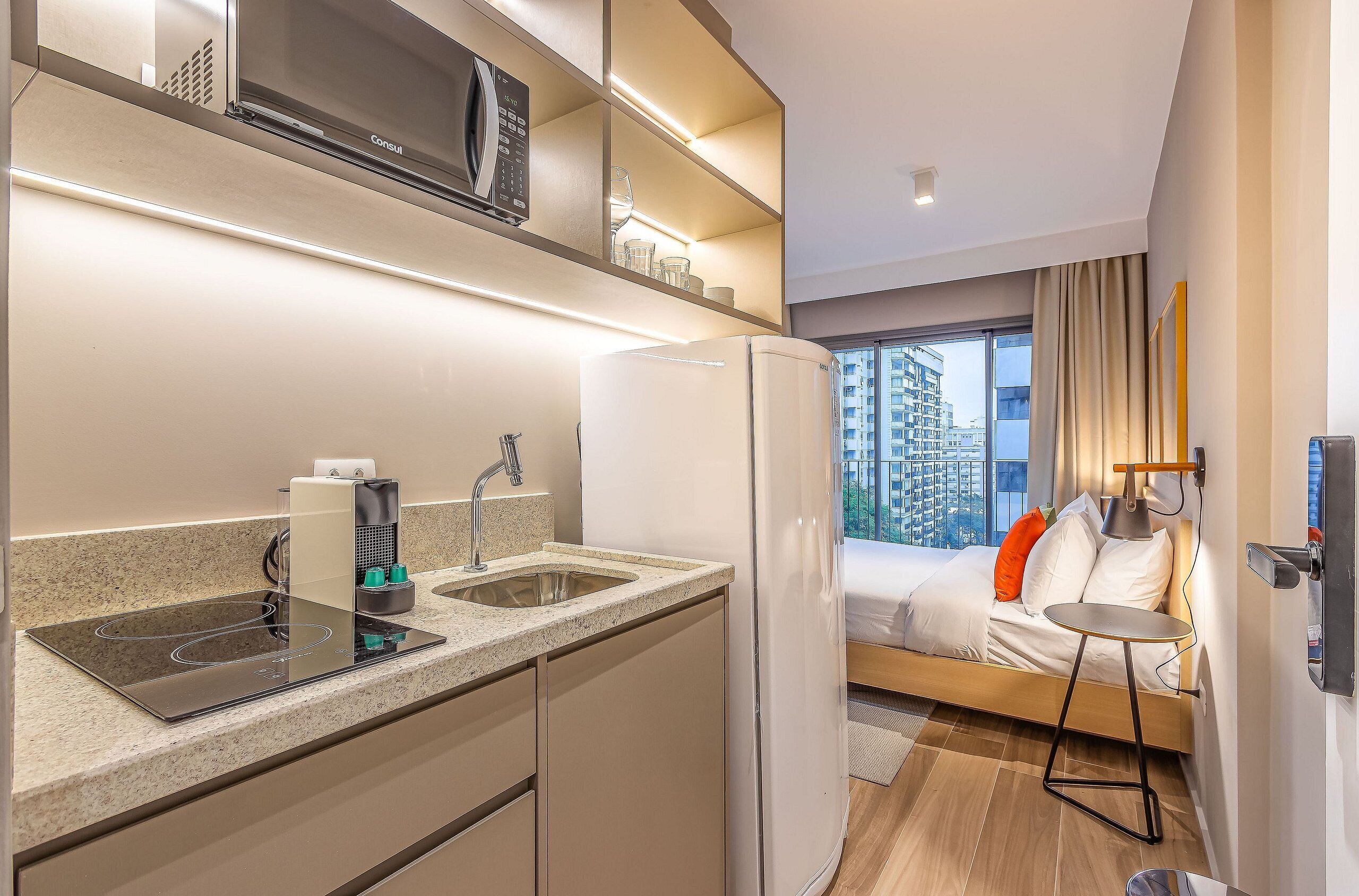 A compact hotel room with a kitchenette, a bed with a view, and a small bedside table.