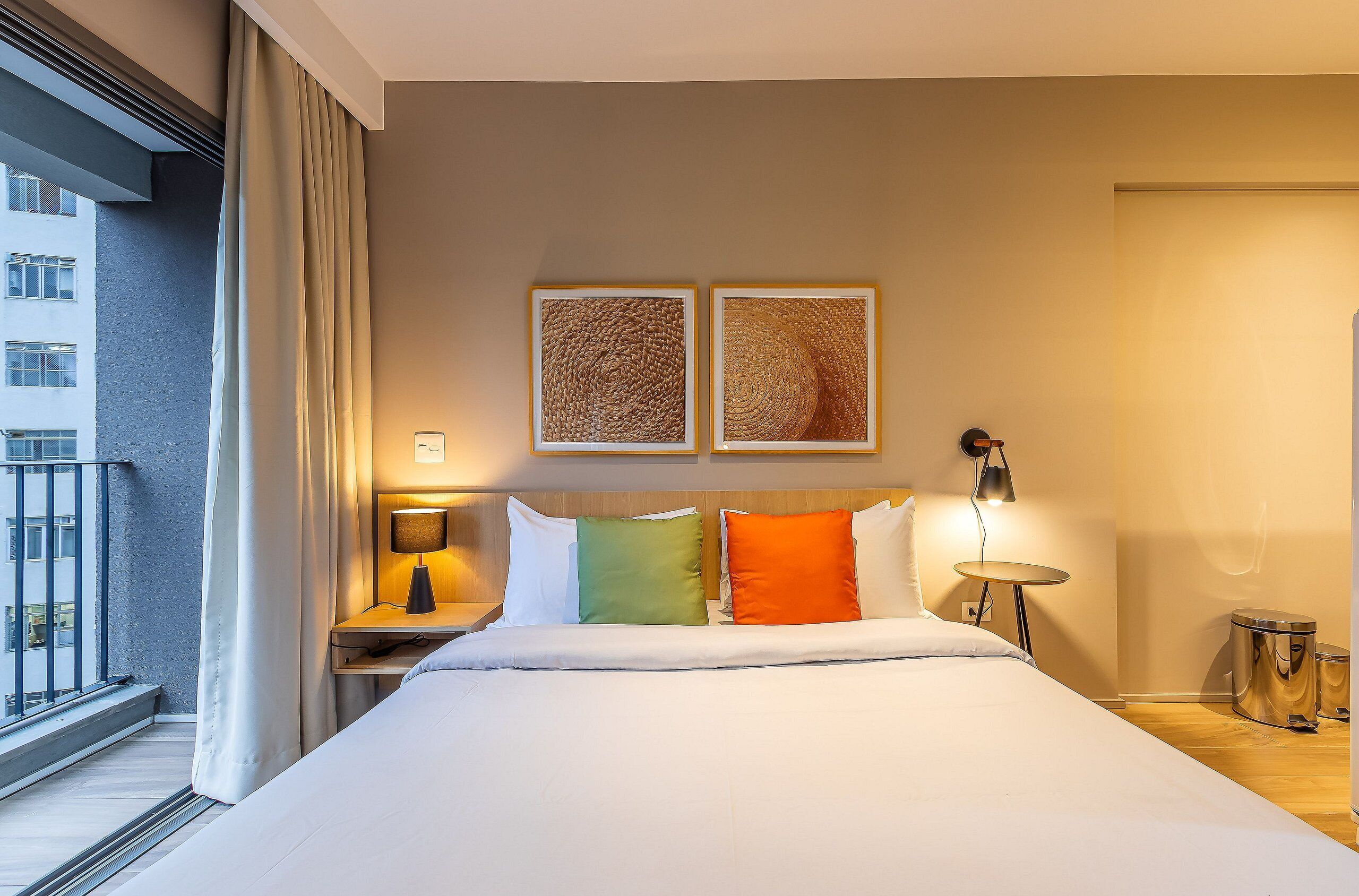 A hotel room with a bed, a nightstand, a lamp, two framed artworks, a window with curtains, and a balcony.
