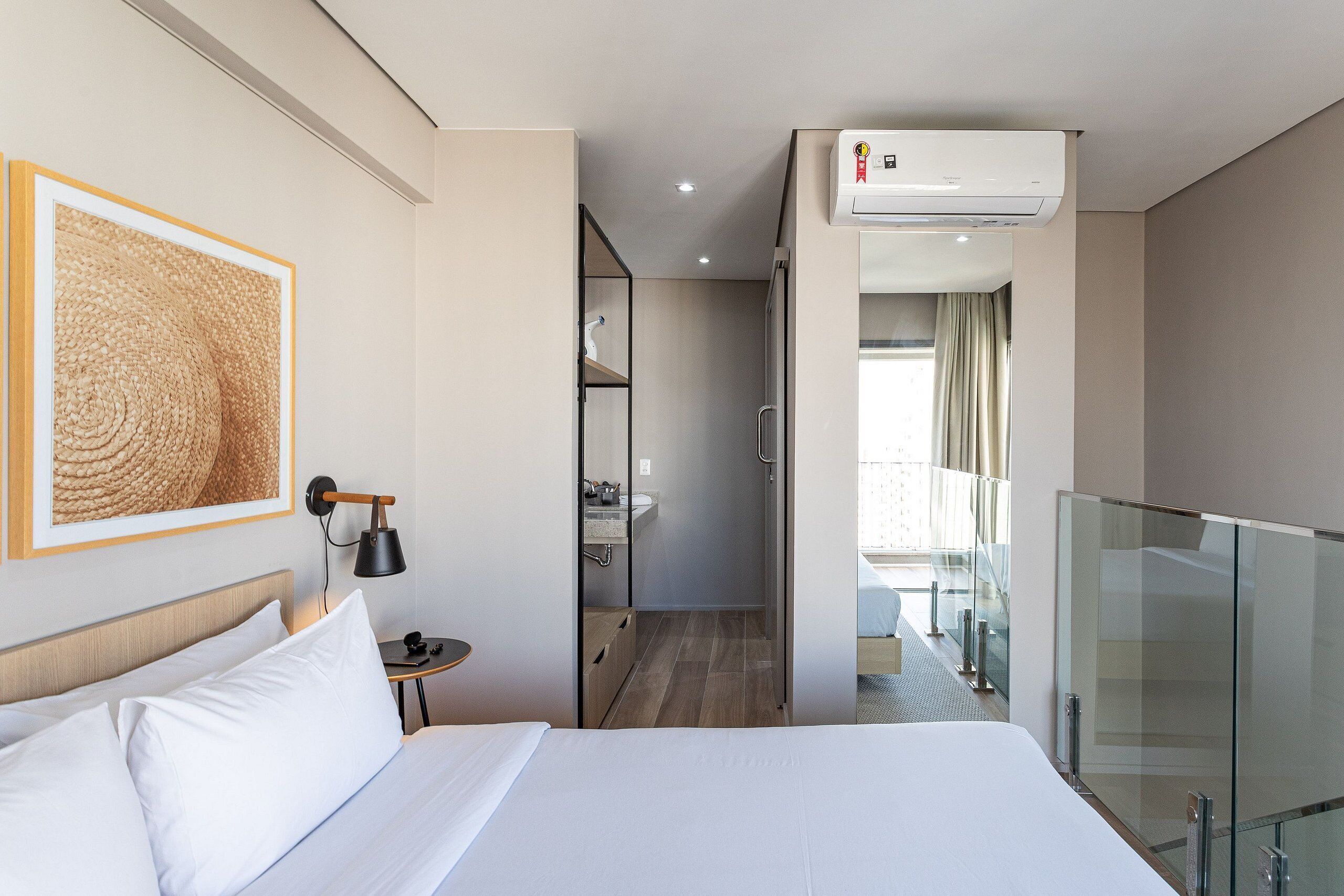 Hotellihuone, jossa on sänky, työpöytä lampun kanssa, kehystetty taideteos, ilmastointilaite ja näkymä parvekkeelle.