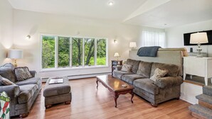 A spacious living room with a large window, a sofa, a coffee table, and a television. - Charming & Scenic Gouldsboro Getaway w/ Patio! (Gouldsboro)
