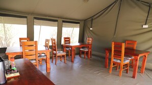 Un espace repas aménagé sous une tente, avec des tables et des chaises en bois, une bouilloire et une bouteille posée sur la table.