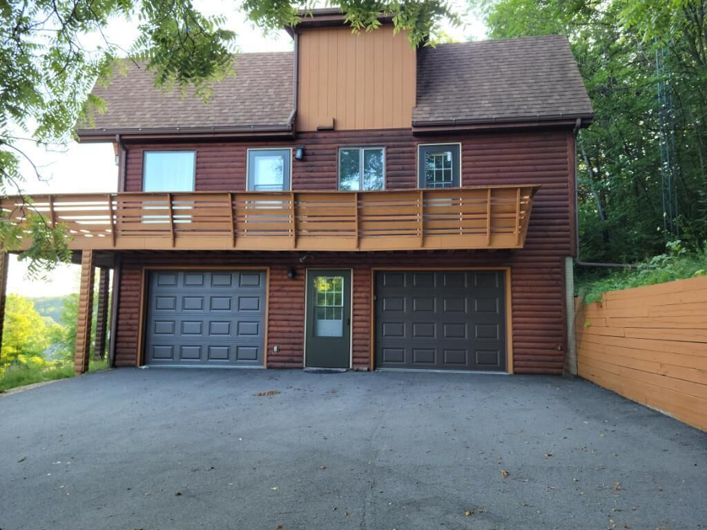 Une cabane en rondins à deux étages, dotée d’un balcon, de deux portes de garage et d’une allée.