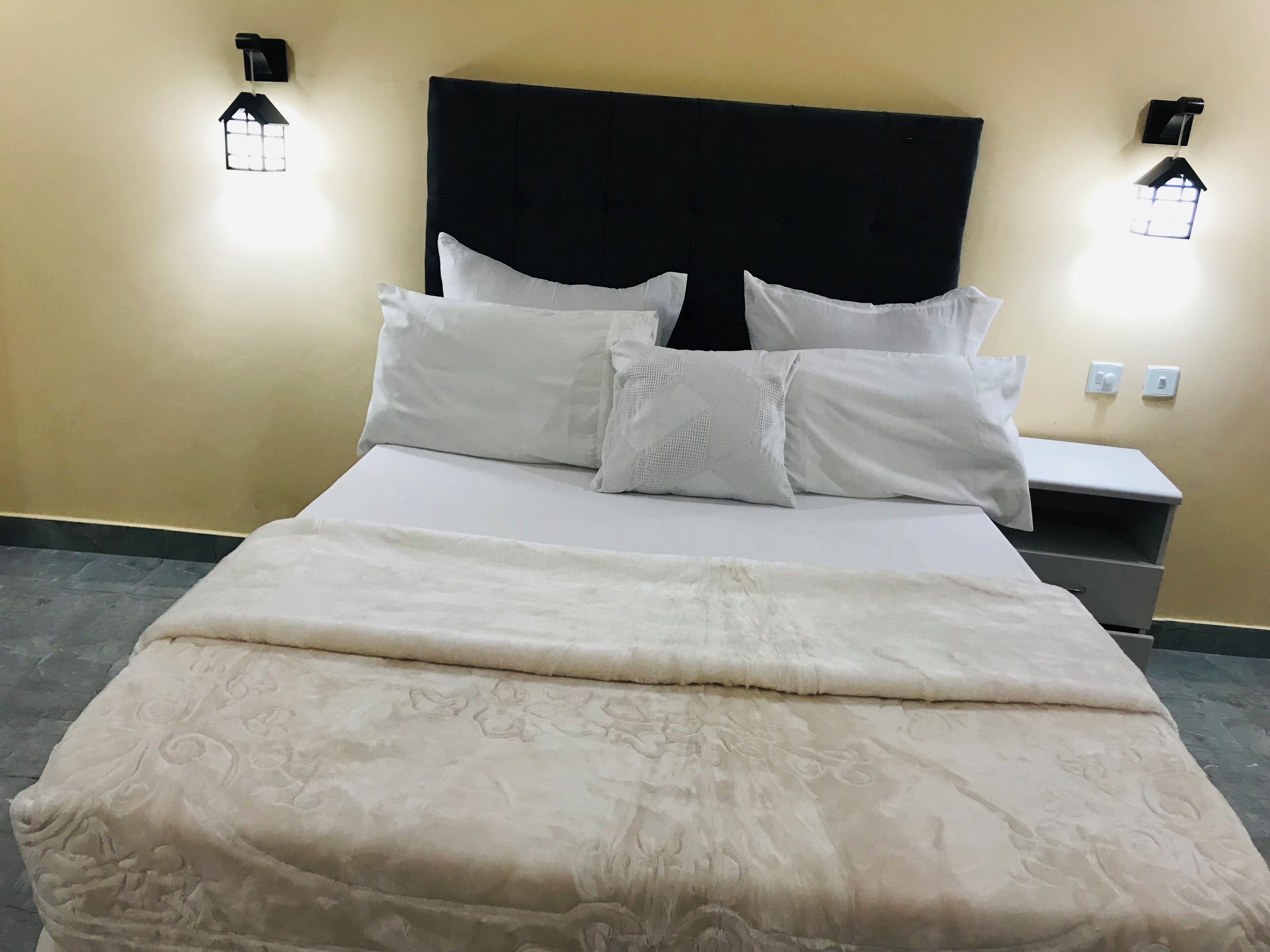 A neatly made bed with a patterned bedspread and white pillows, a dark headboard, and two wall-mounted light fixtures.