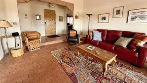 A living room with a red sofa, a wooden coffee table, a patterned rug, a wicker chair, and a lamp. - Vista Gloriosa - panoramic mountain views in Eldorado. (Santa Fe)