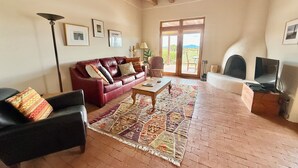 A living room with a red leather sofa, a black armchair, a wooden coffee table, and a patterned rug. There is a television on a wooden stand, a floor lamp, and a large window with a view of the outdoors. - Vista Gloriosa - panoramic mountain views in Eldorado. (Santa Fe)