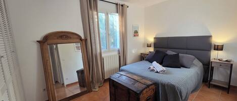 A bedroom with a bed, a wooden dresser, a mirror, a window with curtains, a bedside table with a lamp, and a headboard.