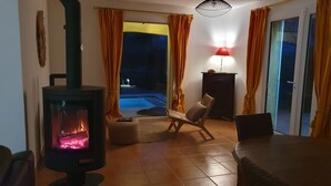Ein gemütliches Wohnzimmer mit Kaminofen, einem bequemen Sessel und Blick auf einen Pool bei Nacht.