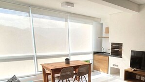 Un salon moderne doté d’une grande fenêtre, un coin repas avec une table et des chaises, une cuisine avec un téléviseur et une porte coulissante donnant sur un balcon.
