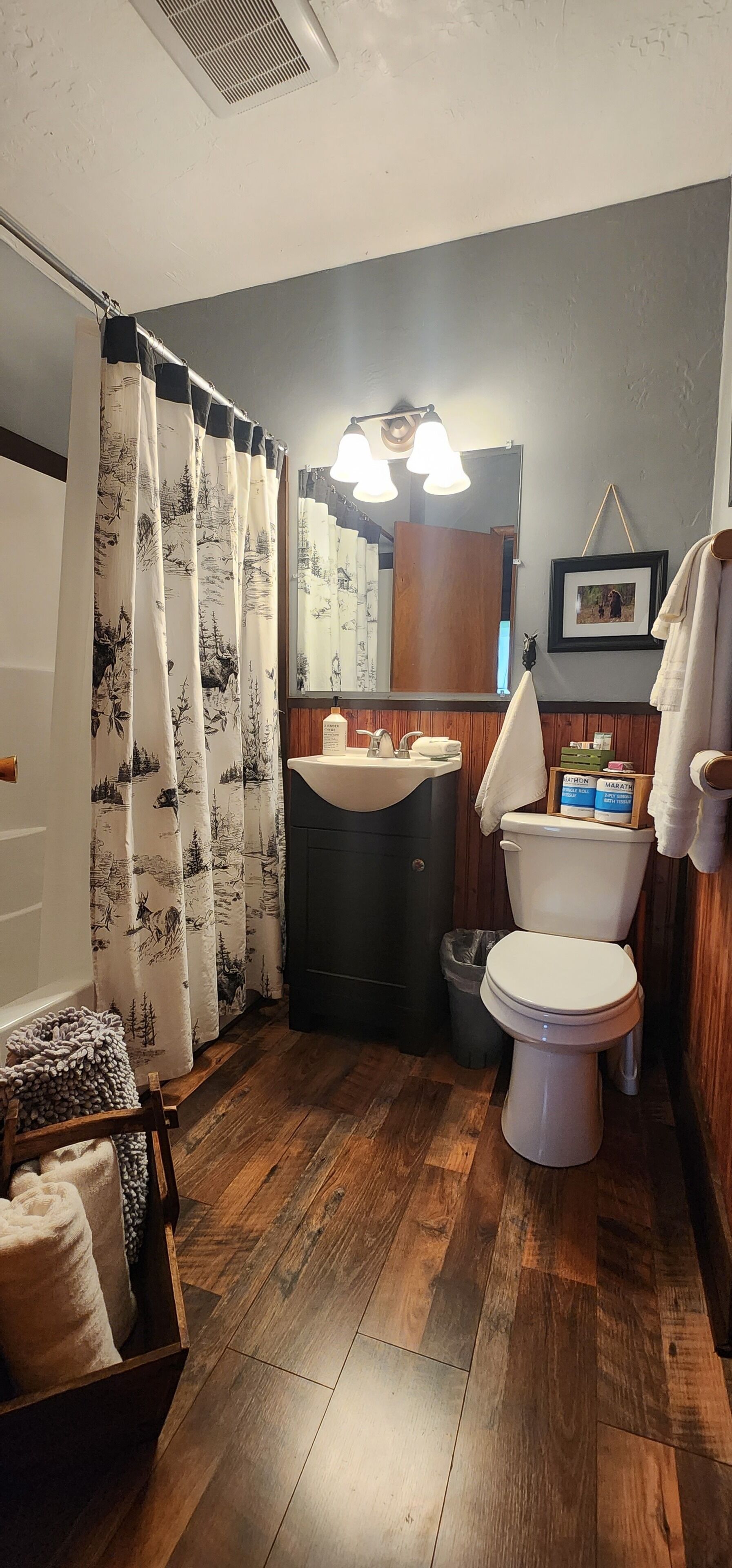 Un baño con suelo de madera, inodoro, lavamanos, espejo y cortina de ducha con motivos naturales.