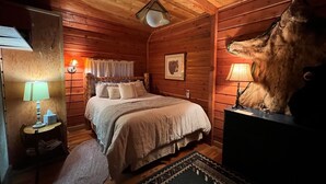 A cozy wooden cabin room with a bed, a nightstand, a lamp, and a bear head decoration. - Bear Creek Guest Ranch (Essex)