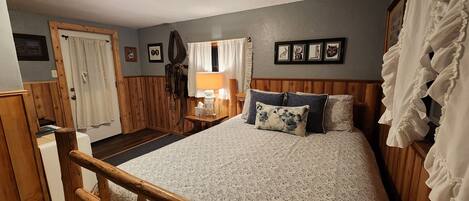 A bedroom with a wooden bed, a nightstand with a lamp, a window with curtains, and a door.
