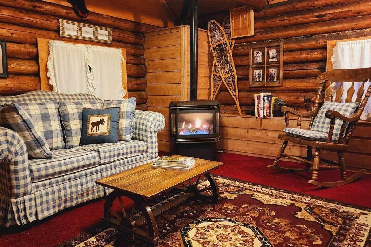 Accogliente interno di una baita in legno con un divano a quadri, una poltrona a dondolo, un tavolino in legno, un camino e decorazioni rustiche alle pareti.