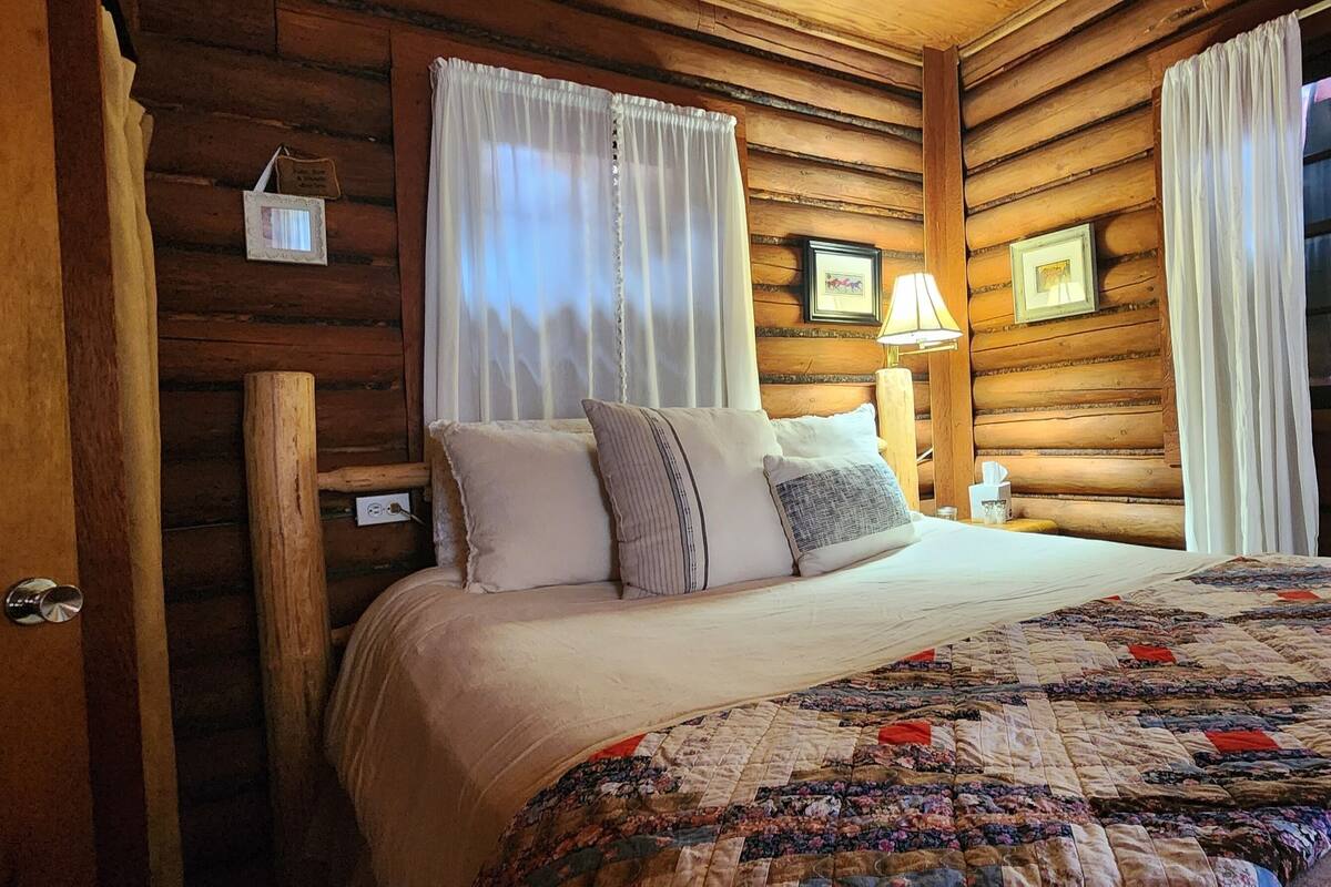 Una camera da letto accogliente in legno con un letto, tende, una lampada e quadri appesi al muro.