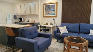 Una sala de estar con un sofá azul, un sillón pequeño, una mesita de mimbre y una zona de cocina con armarios blancos y un cuadro de una playa.