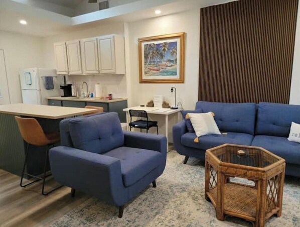 A living room with a blue sofa, a small armchair, a wicker side table, and a kitchen area with white cabinets and a painting of a beach scene.