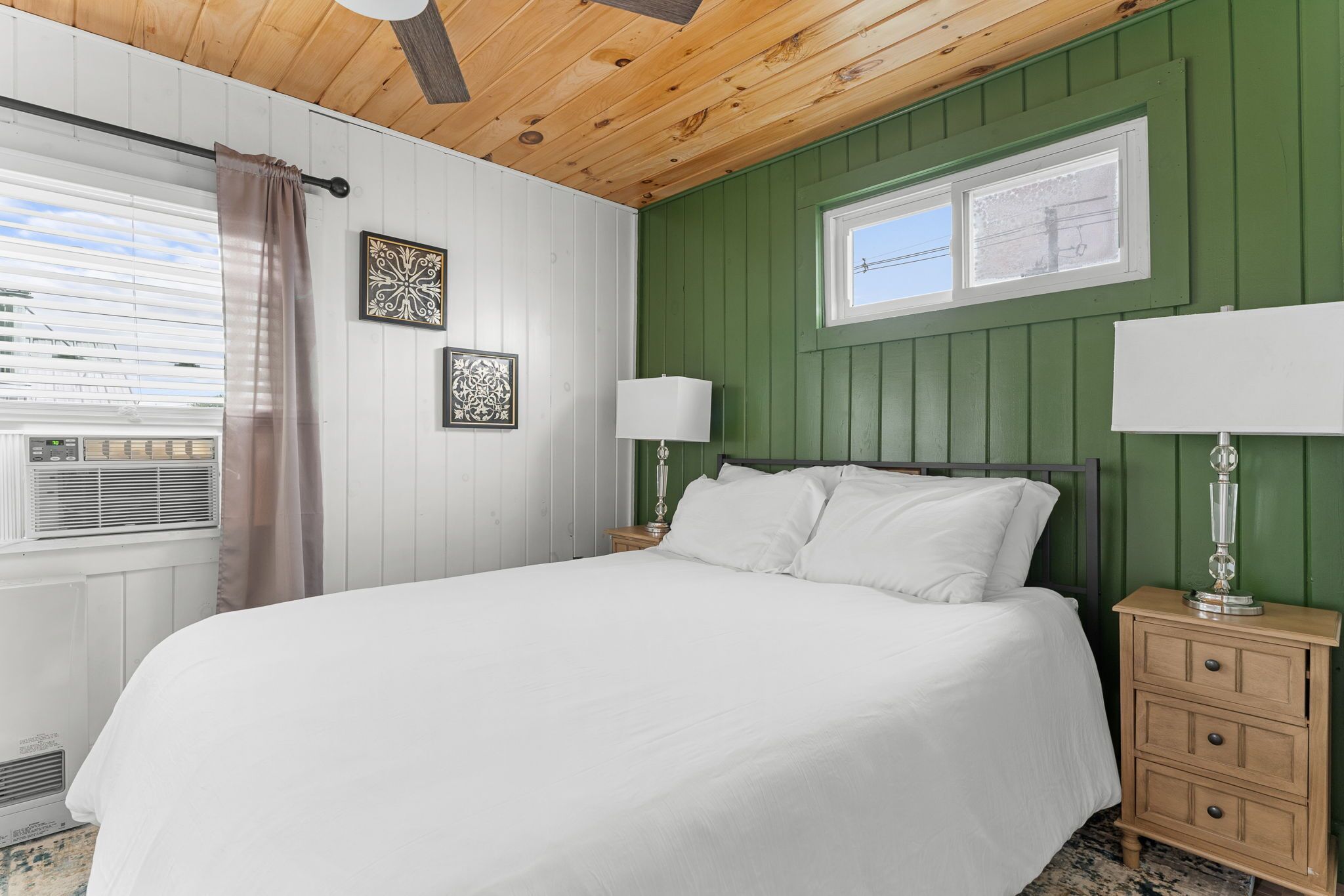A bedroom with a wooden ceiling, green paneled walls, a white bed, two bedside lamps, and a window with blinds.