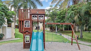 A playground with a wooden playhouse, two slides, and a swing set. - Isla Mazatlán Residence Club (Mazatlán)