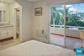 A bedroom with a bed, a bathroom with a toilet, and a sliding glass door leading to a balcony with a view of palm trees. - Isla Mazatlán Residence Club (Mazatlán)