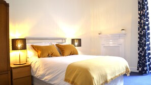 A neatly made bed with a white and beige duvet, two bedside lamps, a wooden nightstand, a white dresser, and a blue patterned curtain. - Charming Spacious Central Apartment (Edinburgh)