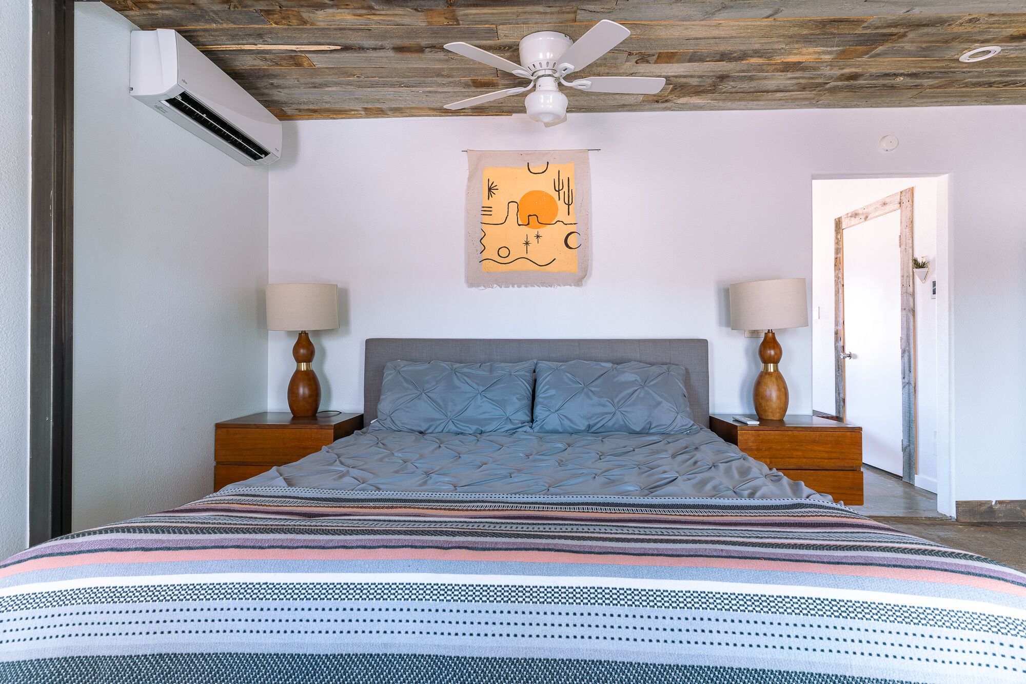 Une chambre à coucher comprenant un lit, deux tables de chevet, un ventilateur de plafond, un appareil de climatisation et une œuvre d’art accrochée au mur.