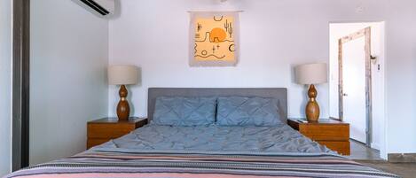 Une chambre à coucher comprenant un lit, deux tables de chevet, un ventilateur de plafond, un appareil de climatisation et une œuvre d’art accrochée au mur.