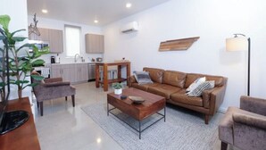 Un salon moderne avec un canapé en cuir marron, une table basse en bois et un tapis gris. On y trouve également un coin repas avec une table et des chaises en bois, et une cuisine à l’arrière-plan.