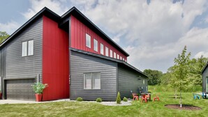 Une maison moderne à deux étages, avec une façade rouge et grise, un garage et un petit patio aménagé avec du mobilier d’extérieur.
