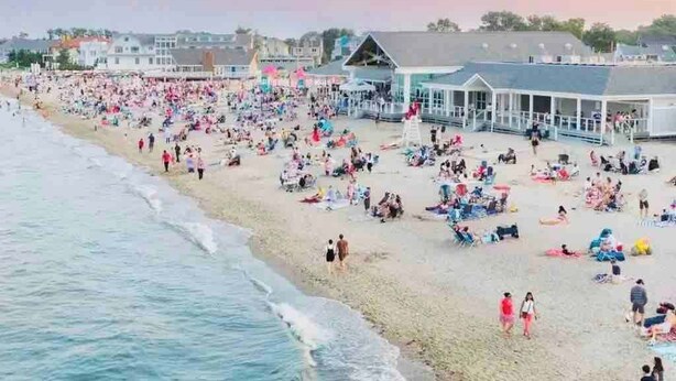 Una playa con gente tomando el sol y caminando por la orilla, un muelle con un área techada y edificios al fondo.