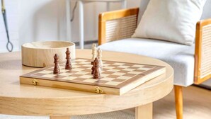 Un tablero de ajedrez de madera sobre una mesa redonda con estampado de cuadros, frente a un salón con sofá y una mesita auxiliar.