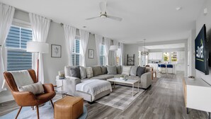 A modern living room with a sofa, armchair, coffee table, and ottoman. There is a television on a stand and a ceiling fan. - Steps to Park Circle! | The Cavalier (North Charleston)