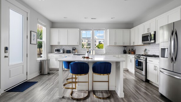 A modern kitchen with white cabinets, stainless steel appliances, and a central island with blue bar stools. - Steps to Park Circle! | The Cavalier (North Charleston)