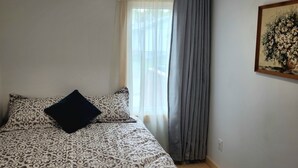Ein Schlafzimmer mit einem Bett, gemusterten Betttextilien, einem schwarzen Kopfkissen und durchscheinenden Vorhängen. An der Wand hängt ein Bild.