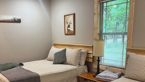 A bedroom with two beds, a ceiling fan, a window with blinds, a framed deer picture, and a bedside lamp.